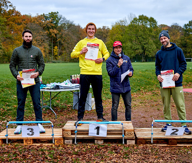 Ein Gewinnerpodest mit drei Plätzen, auf denen jeweils ein Mann steht, während ein Moderator mit einem Mikrofon neben dem Gewinner steht