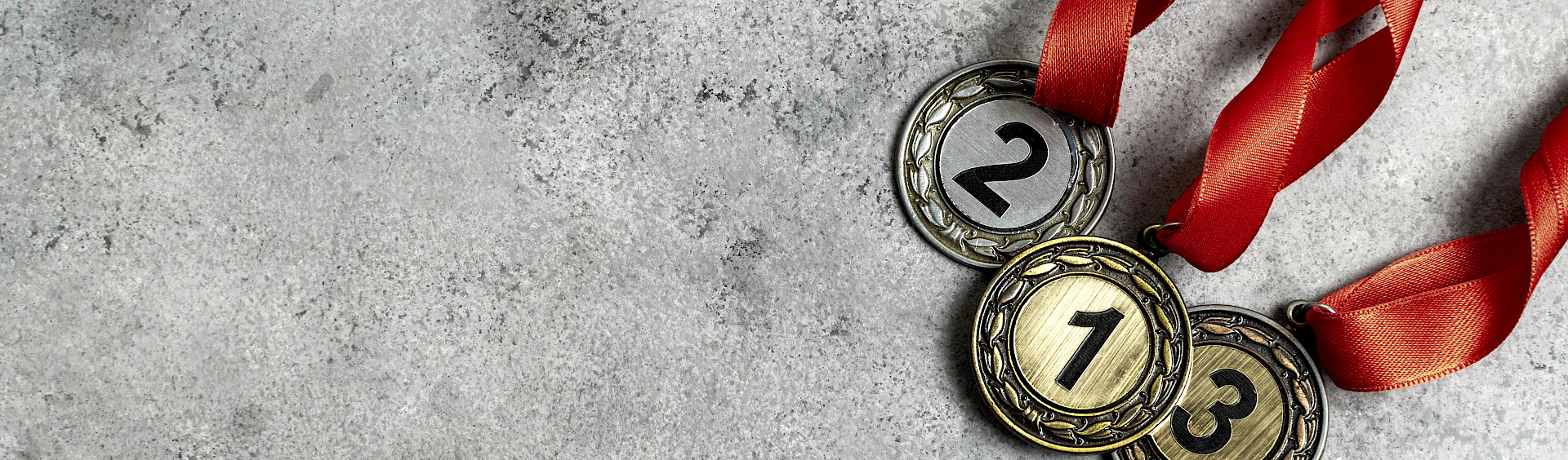 Drei Medaillen mit roten Bändern und den Nummern Eins, Zwei und Drei liegen auf einem grauen Steinhintergund