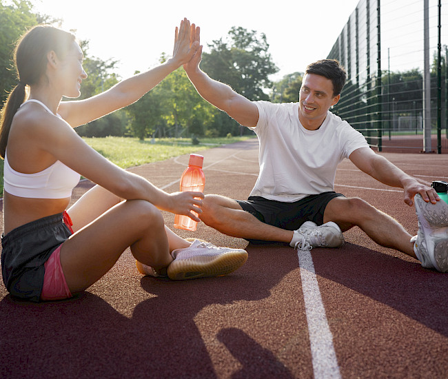Zwei Sportler sitzen mit Wasserflaschen auf einer Rennbahn und klatschen ein