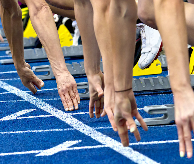 Mehrere Läufer hocken auf einer Laufbahn und bereiten sich auf ein Rennen vor. Man sieht nur die Fingerspitzen, die die Bahn berühren