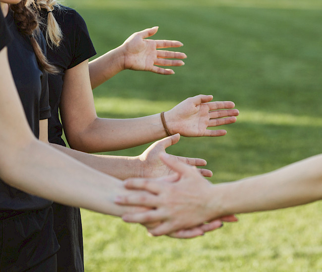 Mehrere junge Sportler stehen auf der linken Seite des Bildes und halten ihre Hände raus, eine Person auf der rechten Seite schlägt bei jedem ein